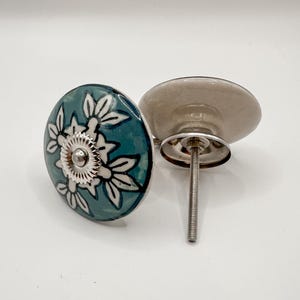 May include: Two decorative cabinet knobs. One knob is teal with a white floral design and a silver metal center. The other knob is a light gray color. Both knobs have a silver metal screw for installation. The knobs are for home decor.
