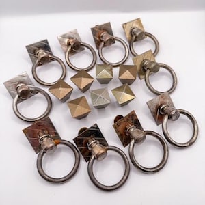 May include: A collection of vintage metal drawer pulls and cabinet knobs. The pulls have square backplates with ring handles. The knobs are geometric shapes in various metallic finishes. The image is on a white background.