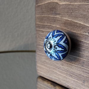 May include: A close-up of a cabinet knob with a floral design. The knob is round with a white base and blue floral pattern. The knob is attached to a wooden cabinet with a natural finish.