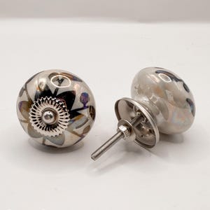 May include: Two decorative cabinet knobs with a floral design. The knobs are primarily silver with black, white, and purple accents. Each knob has a silver metal base and screw for installation. The knobs are likely made of ceramic.