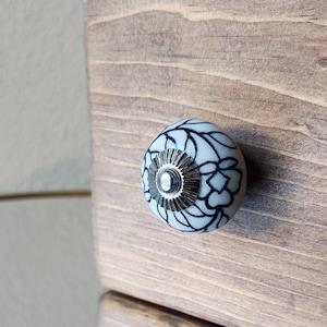 May include: A close-up of a wooden drawer with a decorative knob. The knob is white with a black floral design and a silver metal base. The wood has a natural, light brown finish.
