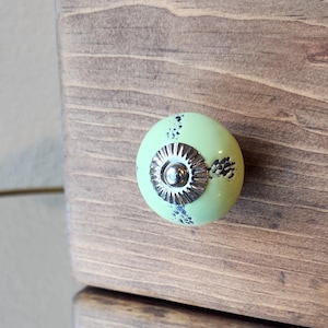 May include: A light green cabinet knob with a silver metal base and a decorative design. The knob is attached to a stained wooden surface. The knob has a round shape and a slightly textured surface.