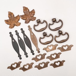 May include: A collection of antique-style cabinet hardware, including leaf-shaped pulls, drawer pulls, and handles in bronze and black finishes. The hardware features heart-shaped cutouts and decorative details.
