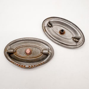 May include: Two antique oval drawer pulls with a distressed silver finish. Each pull features a decorative handle and a central copper-colored rivet. The pulls are designed for furniture restoration or vintage decor projects.