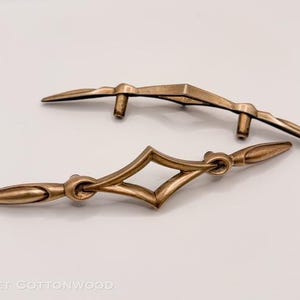May include: Two bronze-colored cabinet pulls. One pull has a diamond-shaped center, while the other has a more angular design. Both have curved handles and are set against a white background. The pulls are likely for furniture.