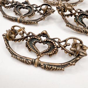 May include: Close-up of four antique bronze drawer pulls. Each pull features a heart-shaped design in the center, surrounded by ornate detailing. The pulls have a vintage aesthetic, with a slightly aged appearance. The text "Sweet Cottonwood" is visible at the bottom.