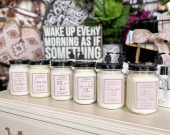 May include: A collection of white jar candles with black lids and pink labels, arranged on a white dresser. The labels feature various phrases. A sign behind the candles reads "WAKE UP EVERY MORNING AS IF SOMETHING".