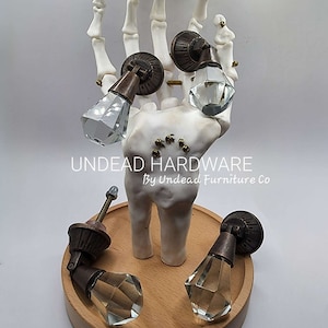 May include: Five antique-style cabinet knobs with clear glass diamond-shaped finials. The knobs are displayed on a wooden base and a skeletal hand.