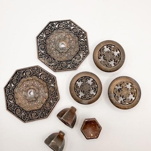 May include: An assortment of antique bronze-toned hardware pieces, including octagonal plates with ornate filigree, circular knobs, and geometric shapes. The pieces are arranged on a white surface, showcasing their intricate designs and aged patina.