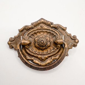 May include: Ornate bronze-colored drawer pull with a central floral design and a curved handle. The pull has an aged appearance, with intricate detailing and a decorative shape. The text "SWEET COTTONWOOD" is at the bottom of the image.