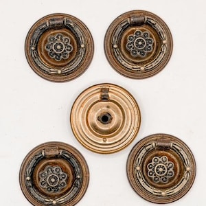 May include: Five antique bronze drawer pulls. Four pulls have a circular design with a central floral motif and a handle. The fifth pull is a circular backplate. The pulls are arranged on a white background.