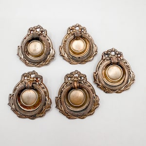May include: Five antique brass drawer pulls with ornate floral detailing. Each pull features a circular handle and a decorative backplate. The pulls have a vintage, aged appearance, with a tarnished finish. The overall design is classic and elegant.