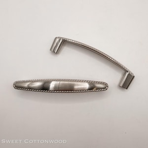 May include: Two brushed nickel cabinet pulls. One is a curved handle with a rope-like border, and the other is a rectangular pull with a similar rope-like border. The pulls are on a white background.