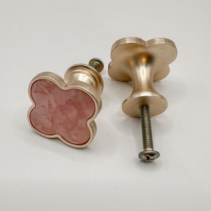 May include: Two decorative cabinet knobs with a cloverleaf design. One knob features a pink marbled center set in a gold-colored frame. The other knob is solid gold-colored. The knobs are for furniture and home decor.