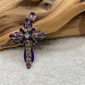 May include: A silver pendant in the shape of a cross, adorned with purple and yellow gemstones. The cross has a purple enamel border and a small purple gemstone at the top. The pendant is set against a wooden background.