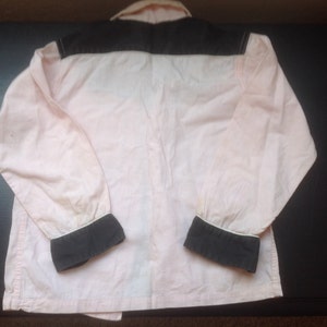 Pu&ograve; includere: Una camicia rosa chiaro a maniche lunghe con carr&egrave; e polsini neri. La camicia ha colletto e polsini bianchi. L'indumento &egrave; realizzato in un materiale morbido, forse cotone. La camicia &egrave; stesa su una superficie scura.