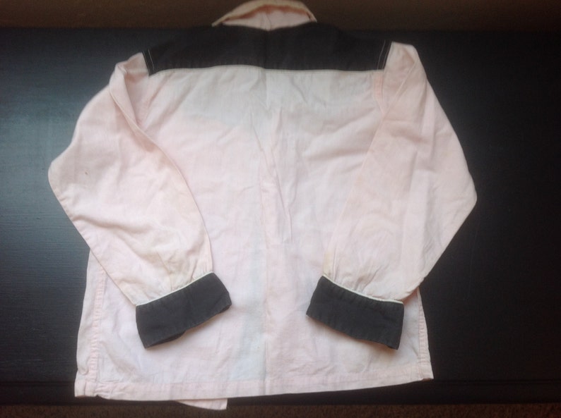 Pu&ograve; includere: Una camicia rosa chiaro a maniche lunghe con carr&egrave; e polsini neri. La camicia ha colletto e polsini bianchi. L'indumento &egrave; realizzato in un materiale morbido, forse cotone. La camicia &egrave; stesa su una superficie scura.