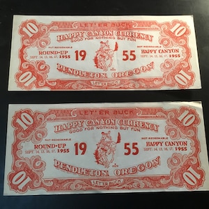 May include: Two vintage "Happy Canyon Currency" bills. Each bill is red and white, featuring text and illustrations. The bills read "Happy Canyon Currency Good for Nothing But Fun" and "Pendleton, Oregon". The year 1955 is prominently displayed.