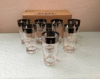 May include: Set of eight clear drinking glasses with a silver rim. The glasses are cylindrical and arranged in a semi-circle in front of a cardboard box. The box has text that reads "GLASS HANDLE WITH CARE".