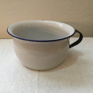 May include: A white enamel chamber pot with a black handle and a blue rim. The pot has a rounded shape and is sitting on a white surface. The interior is white.