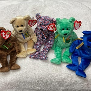 May include: Five colorful plush teddy bears with Ty heart-shaped tags. The bears are brown, tan, patterned, green, and blue. Each bear has a unique design, including ribbons and patches. The bears are sitting on a white surface.