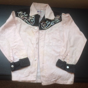 Pu&ograve; includere: Una camicia rosa chiaro con dettagli neri. La camicia ha polsini neri e un colletto nero con ricami a spirale. Bottoni bianchi chiudono la parte anteriore. La camicia &egrave; probabilmente vintage.