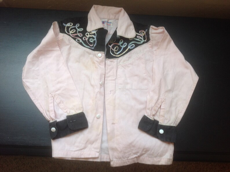 Pu&ograve; includere: Una camicia rosa chiaro con dettagli neri. La camicia ha polsini neri e un colletto nero con ricami a spirale. Bottoni bianchi chiudono la parte anteriore. La camicia &egrave; probabilmente vintage.