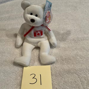 May include: A white plush bear toy with a red ribbon and a Canada flag emblem. The bear has a brown nose and black eyes. A tag is attached to the bear's ear. A yellow sticky note with the number 31 is in the foreground.