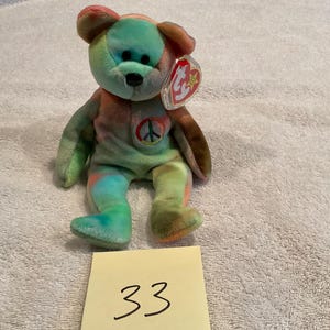 May include: A colorful tie-dye teddy bear plush toy with a peace sign on its chest. The bear has a red, green, and orange color scheme. It has a Ty tag attached. A yellow sticky note with the number 33 is in front of the bear.