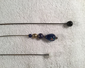 May include: Three hat pins with decorative tops. The top pin has a black, round bead. The middle pin features blue beads, gold accents, and a blue teardrop-shaped bead with floral details. The bottom pin has a white, square bead.