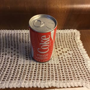 May include: A red and white Coca-Cola can sits on a white, crocheted doily. The can features the Coca-Cola logo in white script. The doily has a delicate, open weave pattern and a scalloped edge. The background is a dark brown surface.