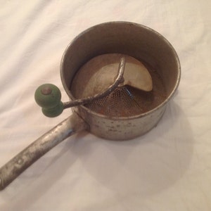 May include: Vintage metal food mill with a perforated bowl, a rotating blade, and a green wooden handle. The handle is attached to a long, sturdy arm. This kitchen tool is designed for processing fruits and vegetables.