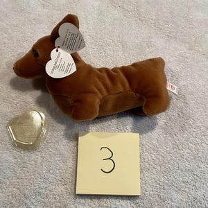 May include: A brown plush dachshund dog toy with heart-shaped tags. The toy is lying on a light gray surface. A small, gold-colored heart-shaped object and a yellow square with the number 3 are also present.