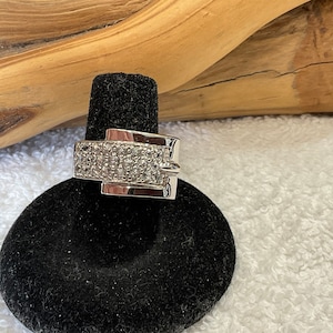 May include: A silver-toned ring with a rectangular design, encrusted with small, clear stones. The ring is displayed on a black ring holder against a neutral background.
