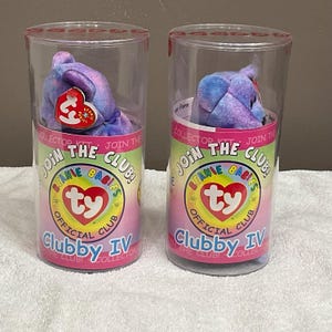 May include: Two Beanie Babies Clubby IV plush toys in clear plastic tubes. The plush toys are purple and blue. The tubes have a pink and green label with the text "Join the Club! Beanie Babies Official Club".