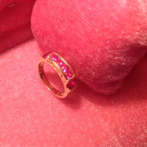May include: A gold ring with a row of bright pink gemstones. The ring is set against a soft, pink background, creating a contrast that highlights the ring's design. The gemstones are evenly spaced.