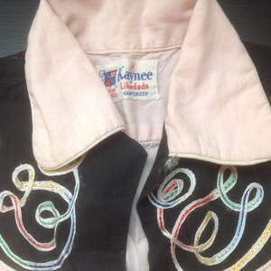 Pu&ograve; includere: Primo piano di una camicia vintage con corpo nero e colletto e polsini rosa chiaro. La camicia presenta vortici ricamati colorati e il nome del marchio "Kaynee Likedads" sull'etichetta.