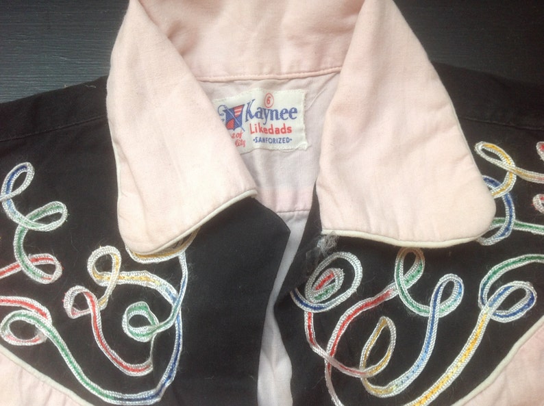 Pu&ograve; includere: Primo piano di una camicia vintage con corpo nero e colletto e polsini rosa chiaro. La camicia presenta vortici ricamati colorati e il nome del marchio "Kaynee Likedads" sull'etichetta.