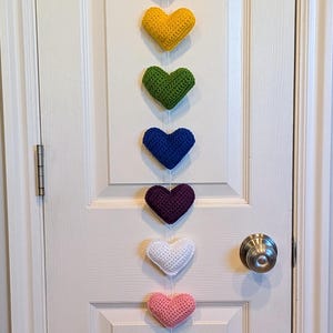 Könnte beinhalten: Eine Kette aus zehn gehäkelten Herzen in Regenbogenfarben, die vertikal an einer weißen Tür hängt. Die Herzen sind rot, orange, gelb, grün, blau, lila, weiß, rosa, türkis und braun.