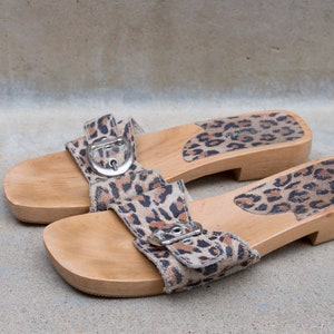 Puede incluir: Un par de sandalias de plataforma de madera con una correa de tela de estampado de leopardo y una hebilla. Las sandalias tienen un acabado de madera natural.