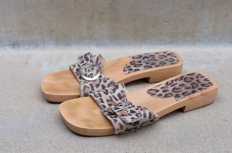 Puede incluir: Un par de sandalias de plataforma de madera con una correa de tela de estampado de leopardo y una hebilla. Las sandalias tienen un acabado de madera natural.