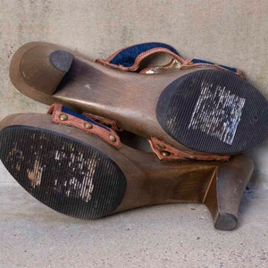 May include: A pair of wooden platform sandals with a denim and brown fabric strap. The sandals have a high heel and a thick platform.