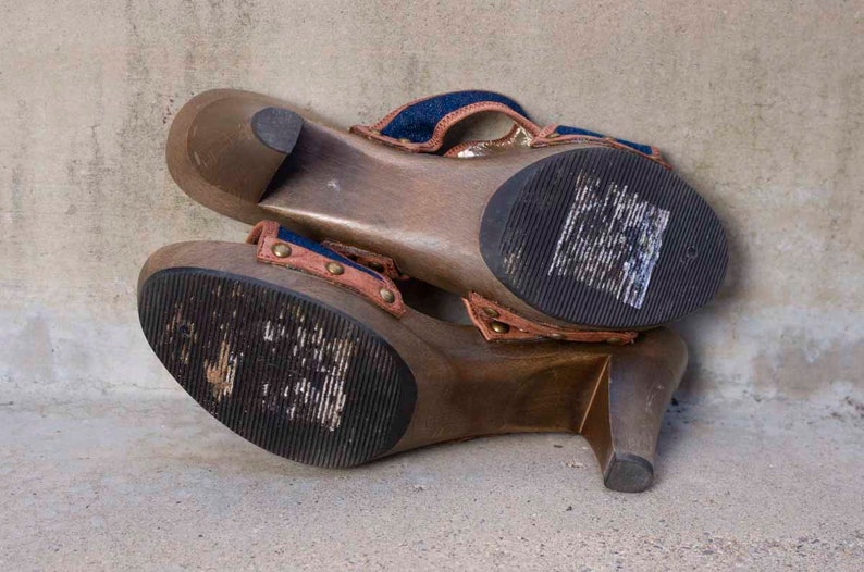 May include: A pair of wooden platform sandals with a denim and brown fabric strap. The sandals have a high heel and a thick platform.
