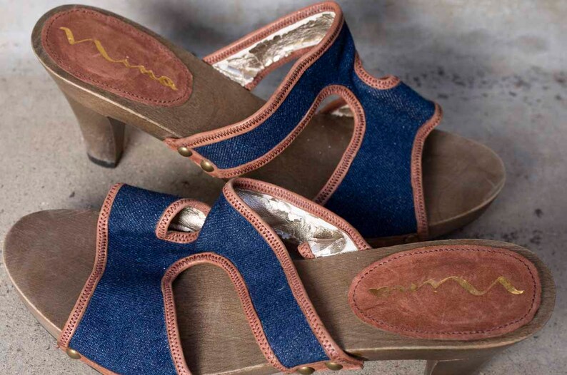 May include: A pair of brown wooden platform sandals with blue denim straps and a gold logo on the insole.