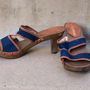 May include: A pair of blue denim and brown leather heeled sandals with wooden platforms. The sandals have a buckle closure and are labeled "Kama"
