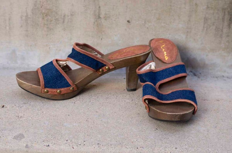 May include: A pair of blue denim and brown leather heeled sandals with wooden platforms. The sandals have a buckle closure and are labeled "Kama"