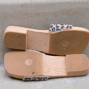Puede incluir: Un par de sandalias de plataforma de madera con una correa de estampado de leopardo. Las sandalias tienen una suela gruesa de madera y una plantilla c&oacute;moda.