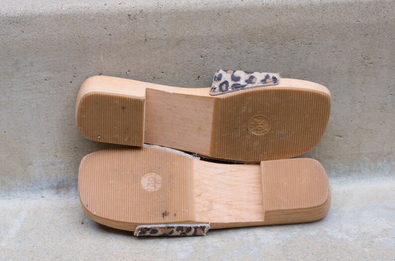 Puede incluir: Un par de sandalias de plataforma de madera con una correa de estampado de leopardo. Las sandalias tienen una suela gruesa de madera y una plantilla c&oacute;moda.