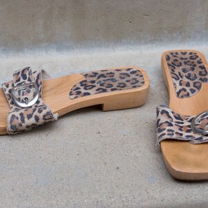 Puede incluir: Un par de sandalias de plataforma de madera con correas de tela de estampado de leopardo y hebillas plateadas. Las sandalias tienen una suela de madera y un tac&oacute;n bajo.
