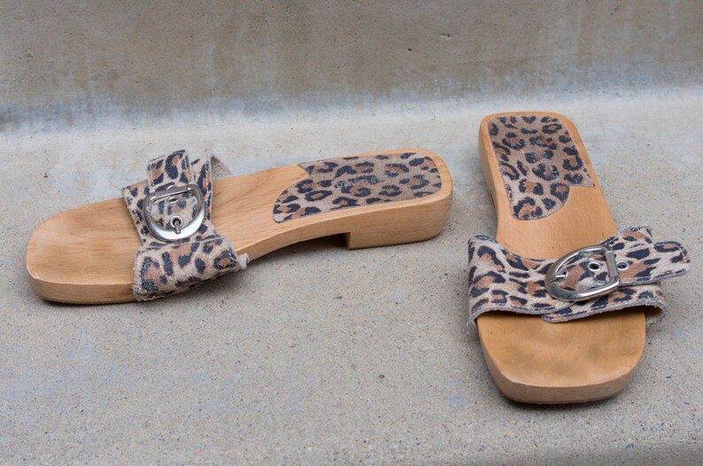Puede incluir: Un par de sandalias de plataforma de madera con correas de tela de estampado de leopardo y hebillas plateadas. Las sandalias tienen una suela de madera y un tac&oacute;n bajo.